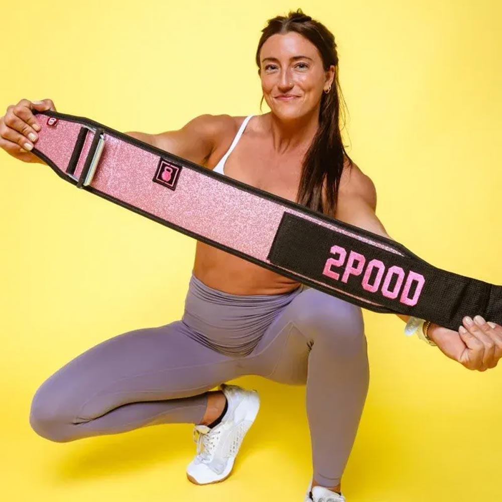 belt-female-gewichtheben Sportlerin in lilafarbener Leggings und weißem Top präsentiert den pinkfarbenen 2POOD Gewichthebergürtel „Bubblegum“ vor gelbem Hintergrund.